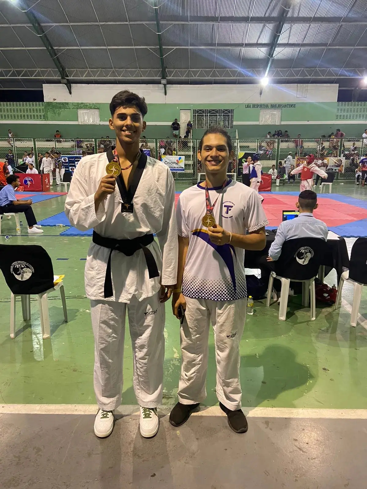 Dois alunos praticantes de taekwondo e participantes do projeto segurando suas medalhas…